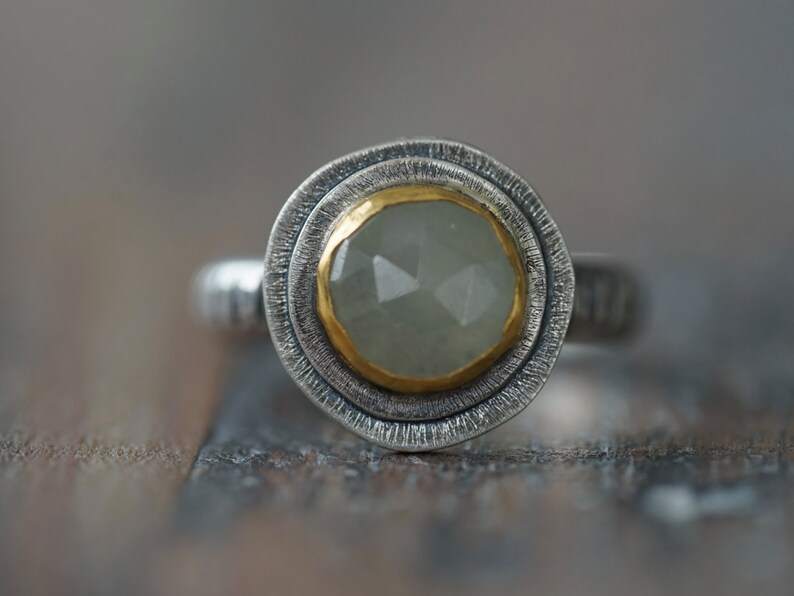 May include: A silver ring with a faceted, pale green gemstone set in a gold bezel. The ring has a textured silver band and a double-layered, textured silver setting around the gemstone.