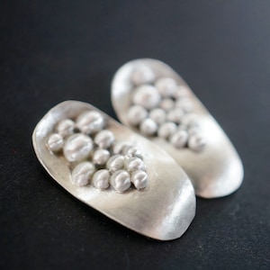 May include: A pair of silver earrings with a textured, oval-shaped design. Each earring features a cluster of small, round, raised bumps in the centre.