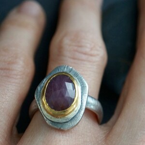 May include: A silver ring with a faceted, oval-shaped, purplish-red gemstone set in a gold bezel. The ring has a textured silver band and a decorative silver setting around the gemstone. The ring is a piece of jewelry.