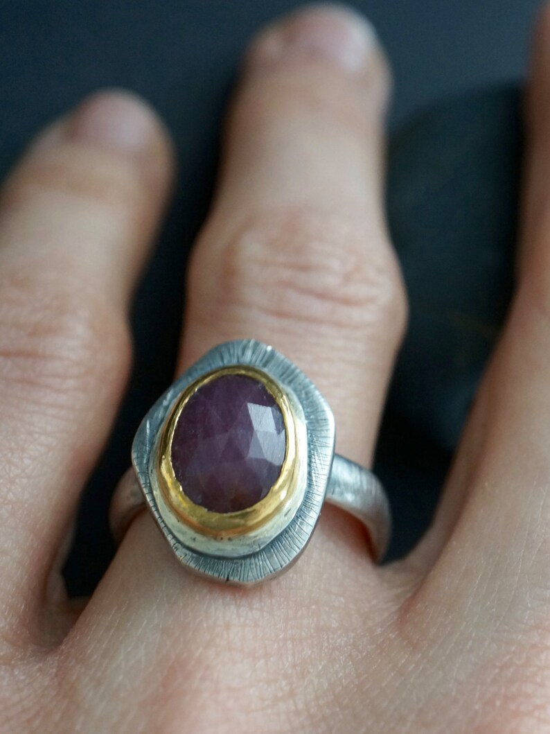 May include: A silver ring with a faceted, oval-shaped, purplish-red gemstone set in a gold bezel. The ring has a textured silver band and a decorative silver setting around the gemstone. The ring is a piece of jewelry.