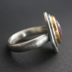 May include: A silver ring with a rectangular, pink gemstone set in a gold bezel. The ring has a textured band and a brushed metal finish, showcasing a blend of textures and colors. The gemstone is a vibrant pink.