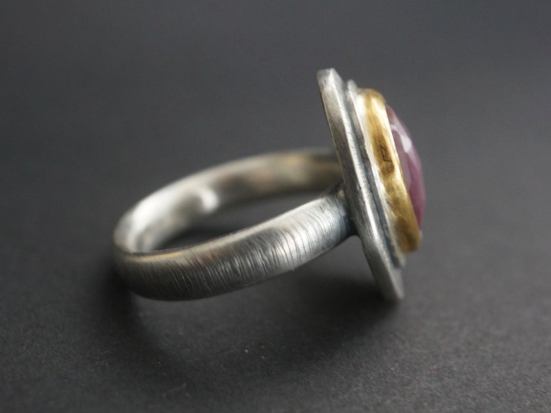 May include: A silver ring with a rectangular, pink gemstone set in a gold bezel. The ring has a textured band and a brushed metal finish, showcasing a blend of textures and colors. The gemstone is a vibrant pink.