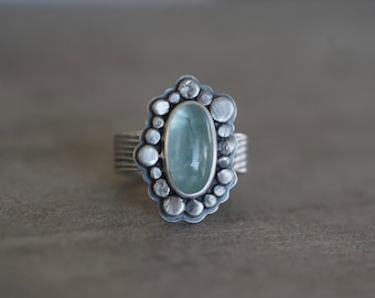 May include: A silver ring with a light blue oval gemstone set in a textured silver bezel. The bezel is surrounded by a ring of small silver beads.