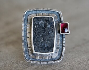 May include: A silver ring with a black stone set in a square frame. A small red gemstone is set on the side of the frame.