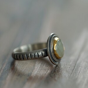 May include: A silver ring with a light green gemstone set in a gold bezel. The ring band has a textured design. The gemstone is oval-shaped and the ring is set against a blurred wooden background.