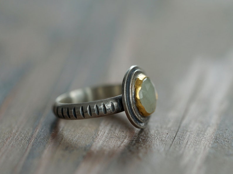 May include: A silver ring with a light green gemstone set in a gold bezel. The ring band has a textured design. The gemstone is oval-shaped and the ring is set against a blurred wooden background.
