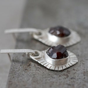 May include: A pair of silver earrings with a dark red gemstone in each. The earrings have a geometric design and a dangling drop.