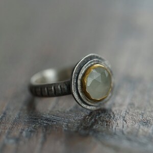 May include: A silver ring with a light green gemstone set in a gold bezel. The ring has a textured band and a double-layered bezel design. The gemstone is round and faceted.