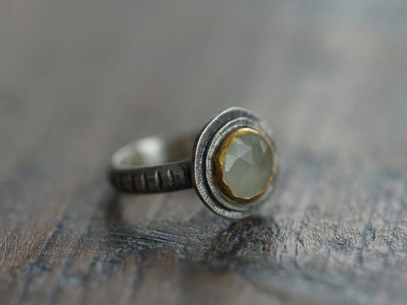 May include: A silver ring with a light green gemstone set in a gold bezel. The ring has a textured band and a double-layered bezel design. The gemstone is round and faceted.