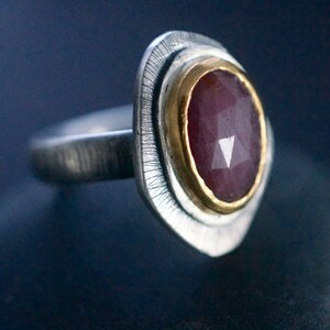 May include: A silver ring with a faceted, oval-shaped, dark red gemstone set in a gold bezel. The ring has a textured silver band and a decorative, textured silver setting around the gemstone.