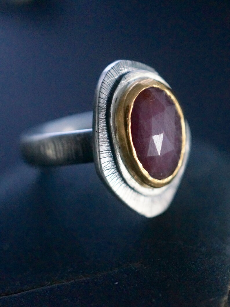 May include: A silver ring with a faceted, oval-shaped, dark red gemstone set in a gold bezel. The ring has a textured silver band and a decorative, textured silver setting around the gemstone.