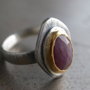 May include: A silver ring with a textured band and a large, faceted, oval-shaped ruby gemstone. The ruby is set in a gold bezel, and the ring has a unique, layered design.