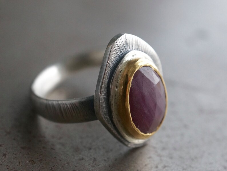 May include: A silver ring with a textured band and a large, faceted, oval-shaped ruby gemstone. The ruby is set in a gold bezel, and the ring has a unique, layered design.