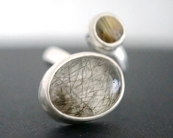 May include: A silver ring with two oval-shaped stones. The larger stone is clear with black inclusions, and the smaller stone is brown with a striated pattern.