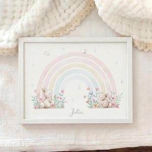 Peut inclure: Une impression d'art encadr&eacute;e en blanc repr&eacute;sentant un arc-en-ciel aux tons pastel au-dessus de deux petits lapins bruns assis dans des parterres de fleurs color&eacute;es. Le pr&eacute;nom Julia est inscrit en lettres cursives au centre, avec des gouttes de pluie.