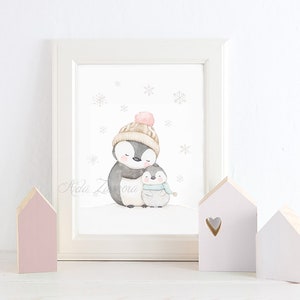 May include: A framed watercolour print featuring a large penguin wearing a beige knit hat with a pink pom-pom and a smaller penguin wearing a light blue scarf. The illustration includes grey snowflakes on a white background with the signature Aida Zamora.