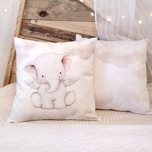 Peut inclure: Deux coussins carr&eacute;s d&eacute;coratifs orn&eacute;s d'une illustration &agrave; l'aquarelle d'un &eacute;l&eacute;phanteau rose p&acirc;le avec de petites ailes, assis sur un nuage. Les coussins pr&eacute;sentent un fond blanc cass&eacute; doux avec des motifs de nuages beige clair.