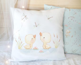 Peut inclure: Un coussin carré blanc orné d'une illustration à l'aquarelle représentant deux canetons jaune clair assis dans un étang bleu avec des roseaux verts, des nénuphars roses et trois libellules volant au-dessus d'eux.