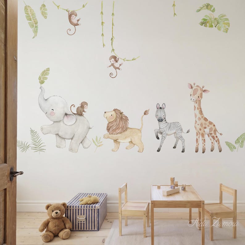 May include: Watercolor wall decals of jungle animals, including an elephant, lion, zebra, and giraffe, with monkeys and foliage accents. The decals are applied to a white wall in a child's room, with a wooden table, chairs, and a teddy bear in the foreground.