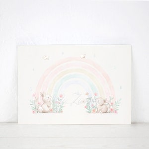 Peut inclure: Une impression d'art rectangulaire pr&eacute;sentant une illustration &agrave; l'aquarelle de deux lapins brun clair assis parmi des fleurs roses et du feuillage vert. Un arc-en-ciel pastel doux surplombe les lapins, avec des gouttes de pluie bleues.