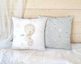 Peut inclure: Deux coussins carrés avec un motif sur le thème de la mer. Un coussin est blanc avec une grande illustration de hippocampe beige et des bulles. Le second coussin est bleu clair avec un motif répétitif de petits poissons, étoiles de mer et bulles.
