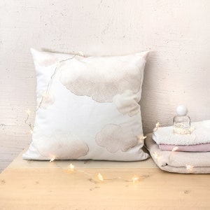 Peut inclure: Un coussin carr&eacute; blanc orn&eacute; d'un imprim&eacute; aquarelle doux repr&eacute;sentant des nuages beiges, drap&eacute; d'une guirlande lumineuse en forme d'&eacute;toiles. &Agrave; c&ocirc;t&eacute; du coussin se trouvent un petit flacon en verre transparent et des serviettes pli&eacute;es.