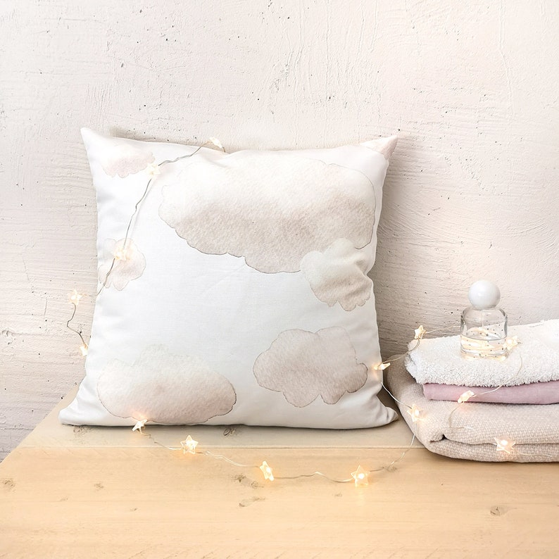 Peut inclure: Un coussin carr&eacute; blanc orn&eacute; d'un imprim&eacute; aquarelle doux repr&eacute;sentant des nuages beiges, drap&eacute; d'une guirlande lumineuse en forme d'&eacute;toiles. &Agrave; c&ocirc;t&eacute; du coussin se trouvent un petit flacon en verre transparent et des serviettes pli&eacute;es.