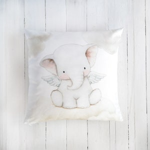 Peut inclure: Un coussin carr&eacute; orn&eacute; d'une illustration &agrave; l'aquarelle d'un &eacute;l&eacute;phanteau blanc avec de petites ailes, assis sur un nuage. L'&eacute;l&eacute;phant a des joues rose p&acirc;le et des yeux sombres, entour&eacute; de nuages beige clair sur un fond blanc.