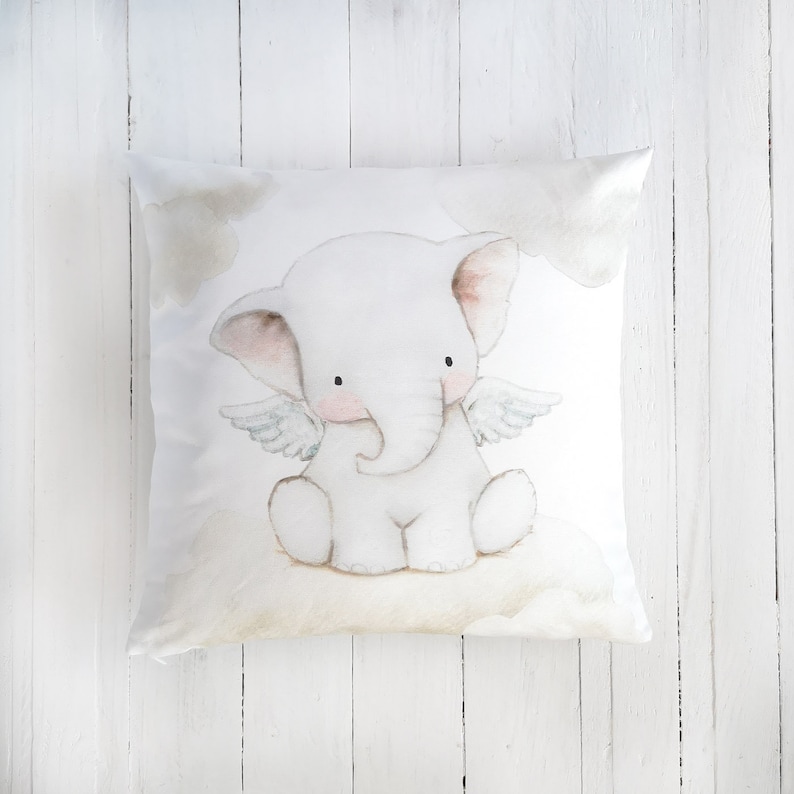 Peut inclure: Un coussin carr&eacute; orn&eacute; d'une illustration &agrave; l'aquarelle d'un &eacute;l&eacute;phanteau blanc avec de petites ailes, assis sur un nuage. L'&eacute;l&eacute;phant a des joues rose p&acirc;le et des yeux sombres, entour&eacute; de nuages beige clair sur un fond blanc.