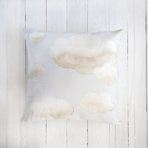 Peut inclure: Un coussin carr&eacute; avec un fond bleu clair orn&eacute; d'un motif de nuages doux de style aquarelle dans des tons beige et brun clair. Les nuages sont dispers&eacute;s sur la surface, variant en taille et en forme, cr&eacute;ant une texture visuelle l&eacute;g&egrave;re.