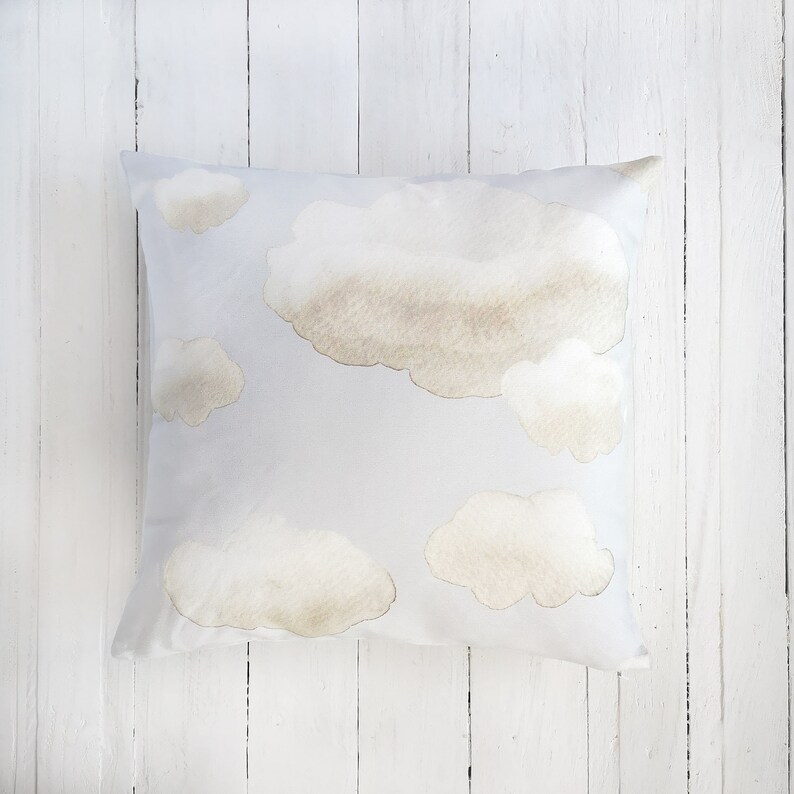 Peut inclure: Un coussin carr&eacute; avec un fond bleu clair orn&eacute; d'un motif de nuages doux de style aquarelle dans des tons beige et brun clair. Les nuages sont dispers&eacute;s sur la surface, variant en taille et en forme, cr&eacute;ant une texture visuelle l&eacute;g&egrave;re.