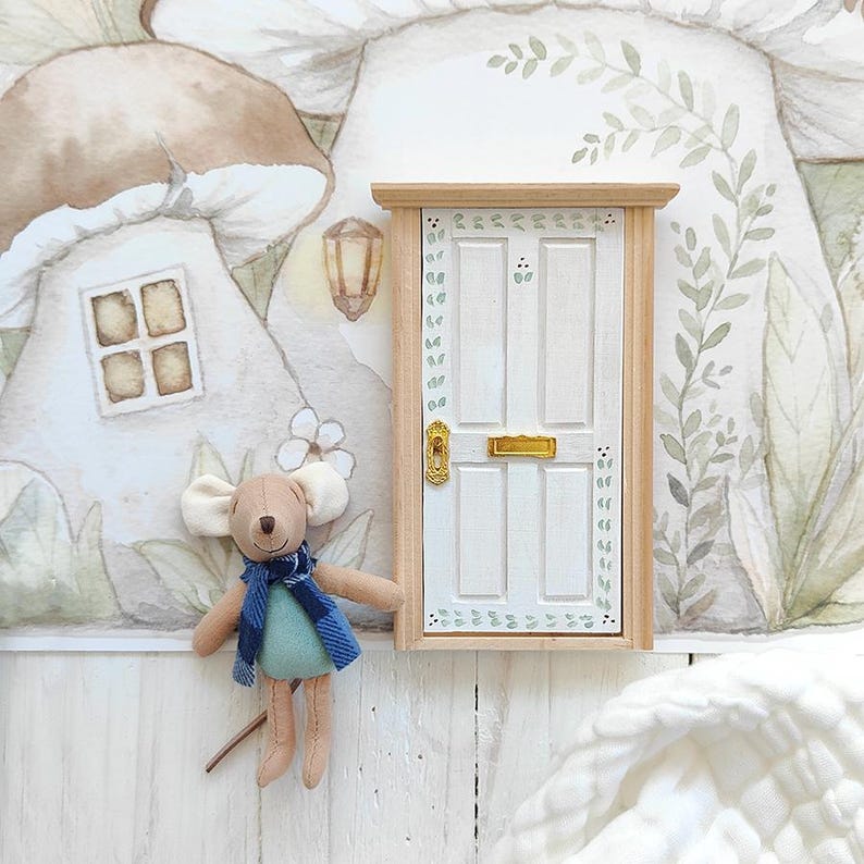 Peut inclure: Une porte miniature en bois blanc avec une poign&eacute;e et une fente &agrave; courrier dor&eacute;es, orn&eacute;e de motifs de feuilles vertes. &Agrave; c&ocirc;t&eacute; de la porte se trouve une petite souris en peluche portant une &eacute;charpe &agrave; carreaux bleus et un haut vert.