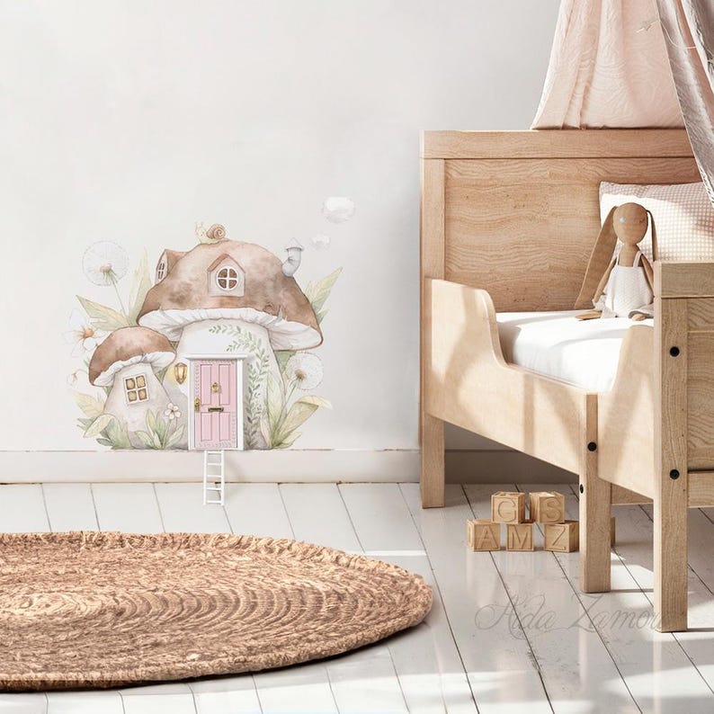 Peut inclure: Un autocollant mural repr&eacute;sentant une illustration &agrave; l'aquarelle de deux maisons champignons brunes avec une porte rose, une petite &eacute;chelle blanche et du feuillage. L'autocollant est pos&eacute; sur un mur &agrave; c&ocirc;t&eacute; d'un lit en bois avec un lapin en peluche.
