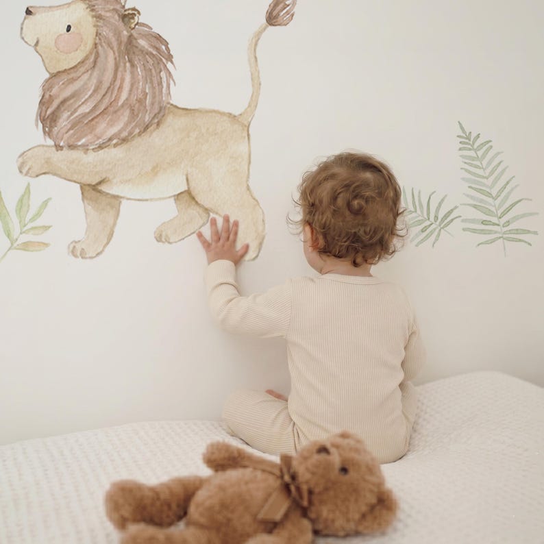May include: A child in cream-colored pajamas sits on a bed, reaching towards a watercolor lion wall decal. A brown teddy bear lies on the bed. Green fern and leaf decals are also on the wall. The lion is tan with a brown mane.