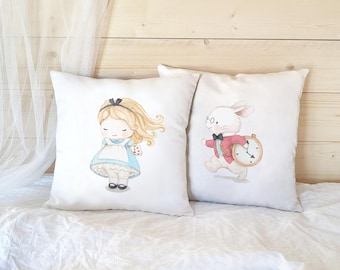Peut inclure: Deux coussins carrés blancs ornés d'illustrations style aquarelle. L'un présente un personnage blond en robe bleue avec un nœud noir, tandis que l'autre montre un lapin blanc portant une veste rouge et un nœud papillon tenant une montre.
