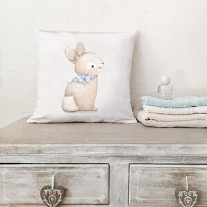 May include: A square white pillow featuring a watercolor illustration of a light brown bunny with a blue bow. The bunny is sitting upright. The pillow is on a wooden surface.