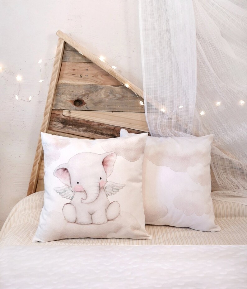 Peut inclure: Deux coussins carr&eacute;s d&eacute;coratifs orn&eacute;s d'une illustration &agrave; l'aquarelle d'un &eacute;l&eacute;phanteau rose p&acirc;le avec de petites ailes, assis sur un nuage. Les coussins pr&eacute;sentent un fond blanc cass&eacute; doux avec des motifs de nuages beige clair.