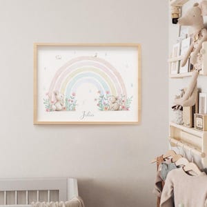 Peut inclure: Une impression d'art murale rectangulaire dans un cadre en bois clair repr&eacute;sentant une illustration &agrave; l'aquarelle d'un arc-en-ciel pastel, deux petits lapins et des fleurs color&eacute;es. Le pr&eacute;nom Julia est &eacute;crit en cursive au bas de l'image.