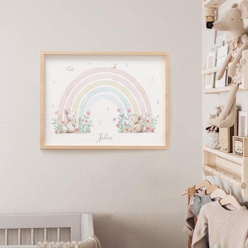 Peut inclure: Une impression d'art murale rectangulaire dans un cadre en bois clair repr&eacute;sentant une illustration &agrave; l'aquarelle d'un arc-en-ciel pastel, deux petits lapins et des fleurs color&eacute;es. Le pr&eacute;nom Julia est &eacute;crit en cursive au bas de l'image.