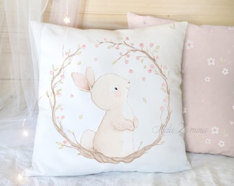 Peut inclure: Un coussin carré blanc orné d'une illustration à l'aquarelle représentant un lapin brun clair debout à l'intérieur d'une couronne circulaire de branches brunes avec de petites fleurs roses et des feuilles vertes. Le texte Alda Zamora est imprimé en cursif.