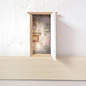 Peut inclure: Une porte miniature en bois avec un cadre blanc et une poign&eacute;e dor&eacute;e. La porte est entrouverte, r&eacute;v&eacute;lant une sc&egrave;ne int&eacute;rieure imprim&eacute;e repr&eacute;sentant une pi&egrave;ce chaleureuse avec une chemin&eacute;e, un fauteuil rose, une biblioth&egrave;que et un tableau.