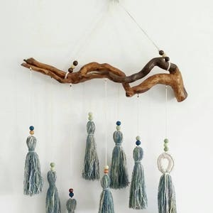May include: A handcrafted wall hanging featuring a natural wood branch with hanging blue and gray yarn tassels. Each tassel is adorned with wooden beads in various colors. The decorative piece is suspended by a thin white cord.