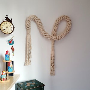 May include: A white macrame wall hanging with a long, knotted rope design and two tassels. The hanging is mounted on a white wall next to a green dresser with floral designs.
