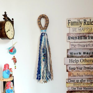May include: A wooden sign with the text "Family Rule BE HAPPY!" and other rules for living a good life. The sign is hanging on a white wall with a blue and white tassel hanging next to it.