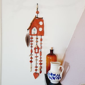 Peut inclure: Une suspension murale en forme de maison en terre cuite avec un cœur et deux cercles. La maison est décorée de perles et d'une plume. La suspension est sur un mur blanc avec un pichet et une bouteille sur une étagère en dessous.
