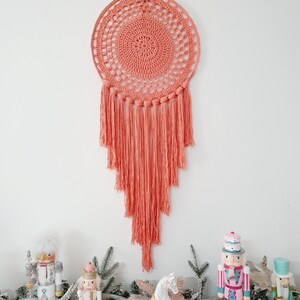May include: A coral-colored, handmade dream catcher with a circular woven design and long fringe hangs on a white wall. Below, festive decorations include toy soldiers and a white horse figurine, creating a holiday theme.