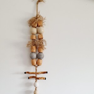 May include: A wall hanging made of natural materials, featuring a variety of wooden beads in beige and gray tones, along with two small pieces of driftwood and a jute tassel.