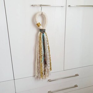 Peut inclure: Un pompon en fil blanc, crème et or suspendu à une porte blanche. Le pompon est noué en boucle et a un accent doré.
