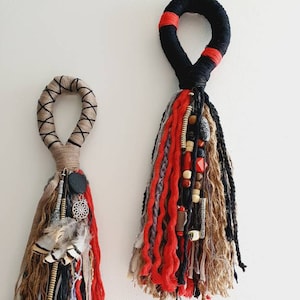 以下が含まれることがあります： タッセルが付いた手作りのマクラメ壁掛けが2つ。大きい方は黒と赤で木製のビーズが付いており、小さい方は茶色と赤で羽根のチャームが付いています。