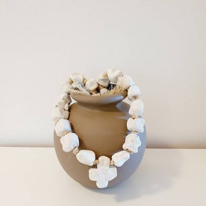 Puede incluir: Una guirnalda de cuentas de cerámica blanca con forma de cruz cuelga de un jarrón de cerámica marrón. La guirnalda está hecha de cuentas individuales conectadas por un cordel.